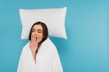 tired woman covered in white duvet standing near flying white pillow and yawning on blue background.