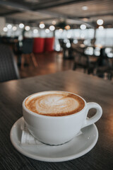 A white ceramic cup with hot coffee drink - cappuccino, americano with milk, latte - stands on a wooden table in a cafe bistro restaurant. Brown foam. Copy space. Selective focus. Breakfast drink.