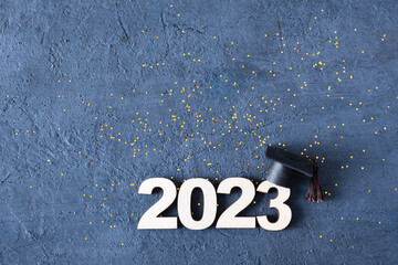 Class of 2023 concept. Wooden number 2023 with graduate hat on dark concrete background with tinsel...