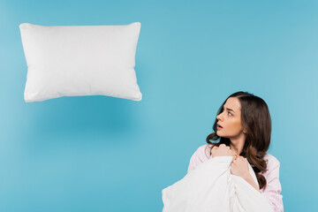 shocked woman in pajamas holding warm duvet looking at levitating pillow on blue.