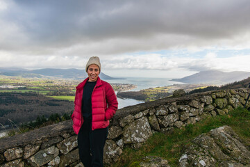 A smiling bi-racial woman hiker wearing a red orange jacket, and white beanie wool hat, with Slieve Gullion, Co. Armagh,  Ring of Gullion, Northern Ireland in the background, showing green hills. - Powered by Adobe