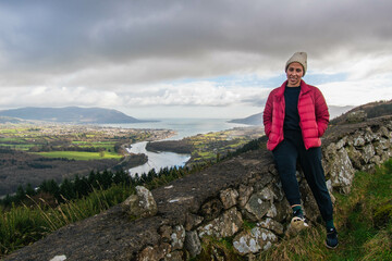 A sitting bi-racial woman hiker wearing a red orange jacket, and white beanie wool hat, with Slieve Gullion, Co. Armagh,  Ring of Gullion, Northern Ireland in the background, showing green hills. - Powered by Adobe