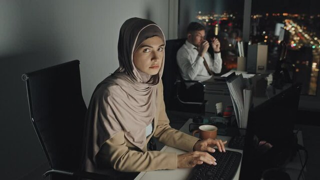 Medium portrait shot of corporate employees working at desks on computers late at night in dark office, then Muslim woman in Islamic hijab turning and looking at camera