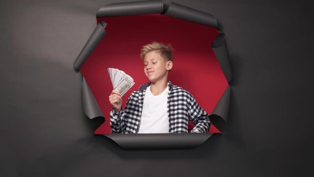 Big money. Rich boy. Wealthy lifestyle. Confident happy teenager kid holding blower of cash dollars posing red background black disrupted paper frame.