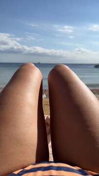 Young beautiful sexy tanned girl in bikini lying on the beach by the sea. View of the sea through the female legs. Vertical video. Striped swimsuit
