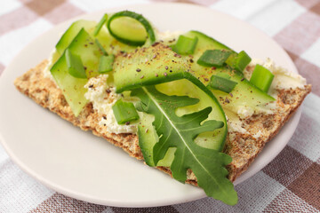 Fresh crunchy crispbread with cream cheese, cucumber, green onion and arugula on checkered tablecloth, closeup
