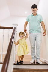 Fototapeta premium Dad holds his daughter's hand and leads her up the stairs. A girl put on her mother's shoes with father on stairs at home. Dangerous situation at home. Kid walking stairs. Baby playing in staircase.