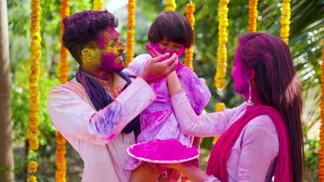 Happy young couple applying holi colors to kid face while looking at camera - concept of holi festival, family bonding and joyful entertainment.