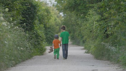 Brothers with back hand in hand walking on empty road, toddler hold a teddy bear