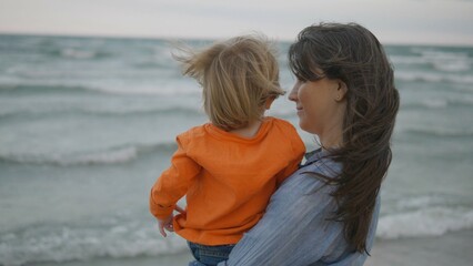 Young mother with back hold baby child look on wild sea waves, empty swing move