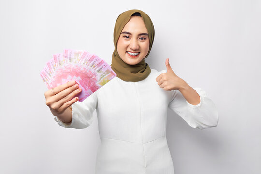 Smiling beautiful young Asian Muslim woman wearing hijab holding cash in rupiah banknotes in hand and showing thumb up gesture isolated on white background. People religious lifestyle concept