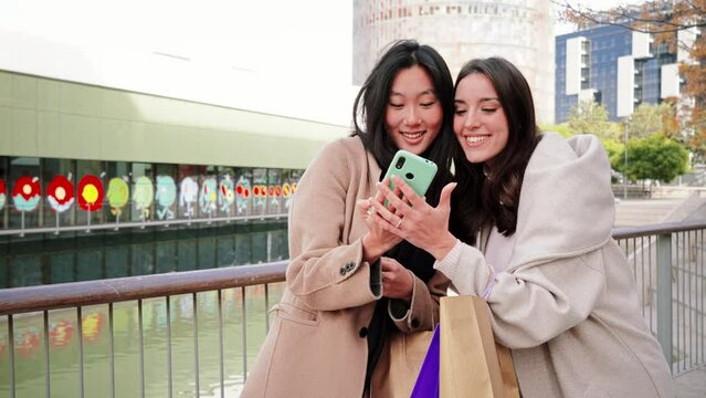 A couple of young women having fun and posing doing a selfie portrait with a cellphone, two girls taking a photo with a smart phone on a shopping day in the city mall. Lifestyle concept. Slow motion
