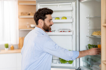 Smiling hungry handsome adult caucasian man with beard takes out food from refrigerator in white...