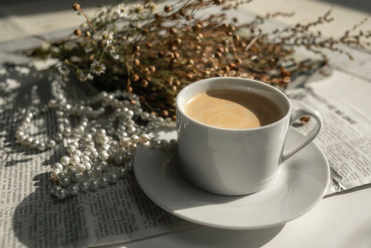 A white cup of coffee on a saucer, with a string of pearls, dried flowers, a magazine and a newspaper. Aesthetic picture, elegant background photo.