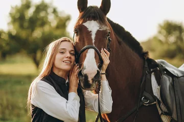 Foto op Canvas Young beautiful female jockey strokes and hugs the horse's head and prepares for the competition. Jumping training in the meadow in summer evening. © sergo321
