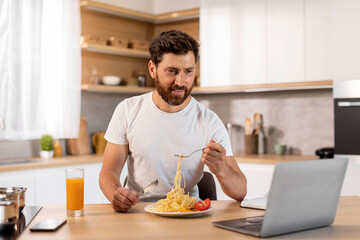 Fototapeta na wymiar Smiling hungry handsome adult caucasian man in white t-shirt watching video on laptop, eating pasta