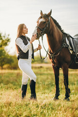 Obraz premium Young beautiful female jockey strokes and hugs the horse's head and prepares for the competition. Jumping training in the meadow in summer evening.