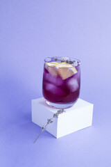 Lavender lemonade with ice and lemon in the drinking glass on the white cube on the purple...