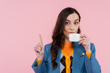 brunette woman in blue blazer drinking coffee and showing wait gesture isolated on pink.