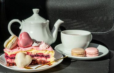 Pink cake with heart shaped macaroons , red tea, white kettle and gold spoon on a black background. Natural light, sunny day