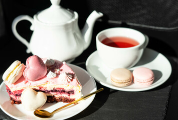 Pink cake with heart shaped macaroons , red tea, white kettle and gold spoon on a black background. Natural light, sunny day
