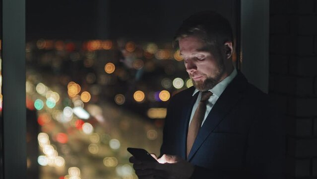 Medium close-up shot of male executive in suit standing by window in dark office at night, typing emails on smartphone, rubbing forehead and eyes, with shimmering bokeh streetlights in background