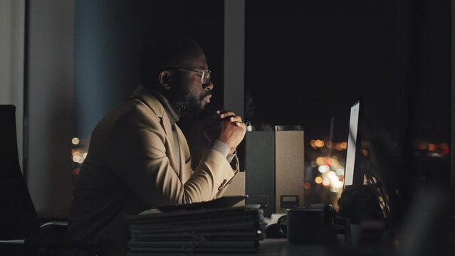 Waist-up shot of African American manager working on computer at midnight alone in dark office, then removing glasses and rubbing eyes and face by hand, with blurred city lights behind window