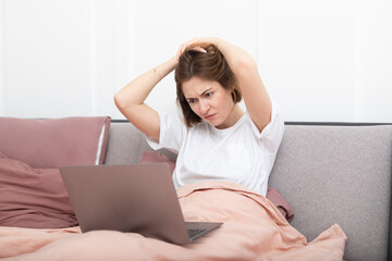 Shocked woman with hands on head working on laptop in the bed. Surprised and upset girl looking at her computer in the bedroom