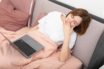 Tired woman lying on bed and working on laptop. Yawning girl looking at the screen of notebook in the bedroom