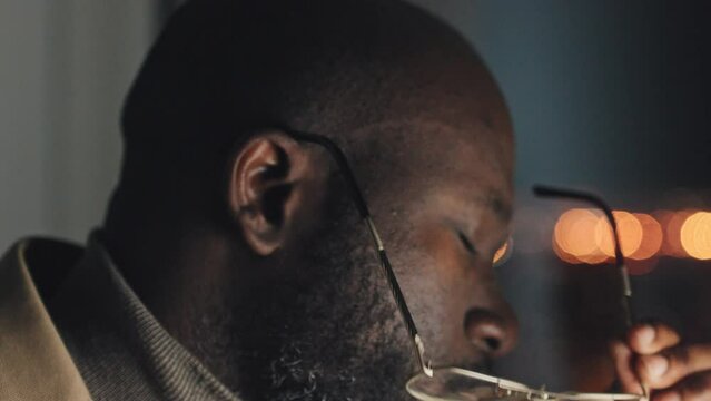 Close-up shot of face of exhausted bald African American man with beard and moustache looking at computer, then taking off glasses and rubbing his eyes, with bokeh lights in dark background