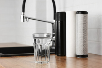 A glass of clean fresh water and set of filter cartridges on wooden table in a kitchen interior. Installation of reverse osmosis water purification system. Concept Household filtration system. - obrazy, fototapety, plakaty