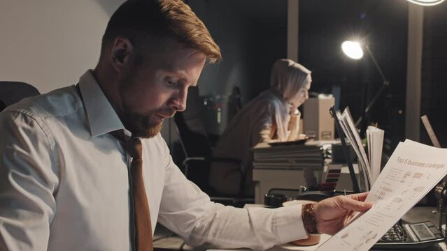 Medium close-up shot of male corporate employee sitting at desk and working in dark office at night, looking through and reading financial documents with graphs, woman in hijab in background
