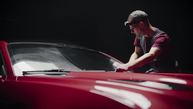Automotive Detailer Polishing a Car Windshield with a Microfiber Towel. Specialist Wearing a Headband Lamp for Better Visual Aid for Spreading a Chemical Wax Solution Against Rain. Detailing Shop