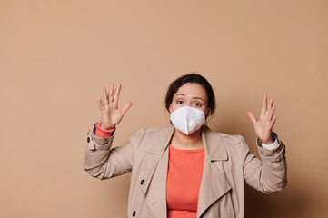 Shocked middle-aged multi-ethnic woman in medical mask, gesturing, expressing stupefaction looking at camera, isolated beige background. Quarantine, self isolation, social distance concept. Copy space