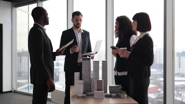 Multiethnic real estate agents, standing near table with 3d city model in office with panoramic city view, while handsome male project manager presenting them report about company's activity on laptop