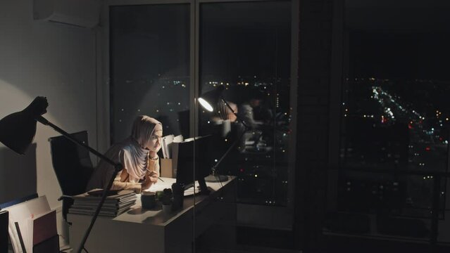Full shot of female office worker in Muslim hijab sitting alone at desk by window late in evening, reading documents on computer and studying paperwork, with dark nighttime cityscape in background