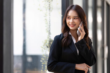 Happy young asian business woman wearing suit holding mobile phone standing in her workstation office
