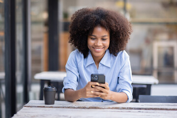 Smiling young woman sitting on chair holding mobile phone using cellphone device, looking at...