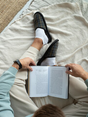 Male feet in white socks and gray plaid slippers on the bed. A man reads a book sitting on couch....