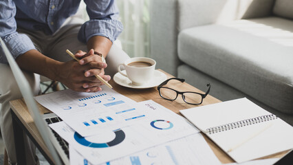 Businessman accountant setting at working space with laptop and holding a pen and taking notes in a notebook and checking financial reports from documents lying on his desk at home office.
