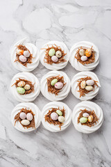 Easter treat - set of white meringues in shape of nest with multicolored candy chocolate eggs over marble background. Overhead view, flat lay, copy space. Holiday symbol