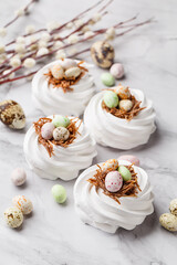 Fototapeta na wymiar Easter treat - set of white meringues in shape of nest with multicolored candy chocolate eggs, quail eggs and pussy willow twigs over marble background. Side view. Holiday symbol