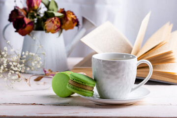 French macaroon dessert. Two green macaroons and a white cup of coffee on a white wooden table against the backdrop of an open book and a winter bouquet of dried flowers