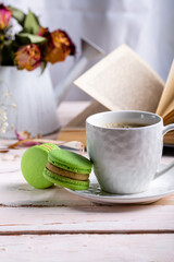 French macaroon dessert. Two green macaroons and a white cup of coffee on a white wooden table against the backdrop of an open book and a winter bouquet of dried flowers