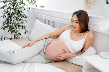 Obraz na płótnie Canvas A pregnant woman with a big open belly on a bed at home dreams of having a baby. The expectant mother is waiting and preparing for the birth of a child in a bright bedroom