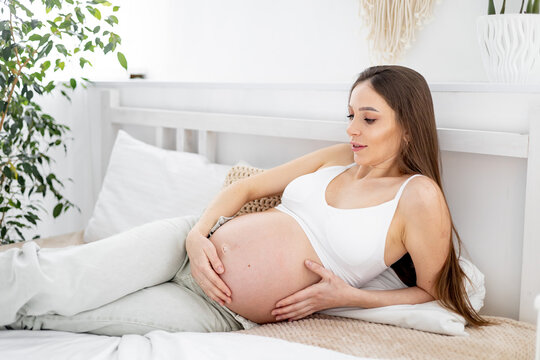 A pregnant woman with a big open belly on a bed at home dreams of having a baby. The expectant mother is waiting and preparing for the birth of a child in a bright bedroom