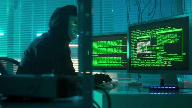 Hacker in a white mask is typing on a computer keyboard and cracking a password. Access denied, access granted. A man in a black hoodie and gloves. The mask reflects green symbols of program codes.