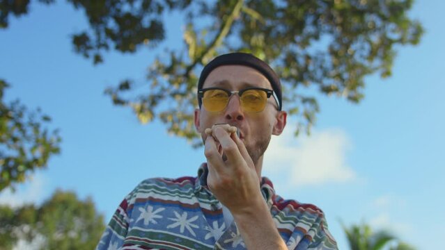 Portrait of man smoking marijuana from pipe. Male hipster in yellow sunglasses smokes weed and blows fume into camera. Cannabis smoker person outdoor with tropical trees at background