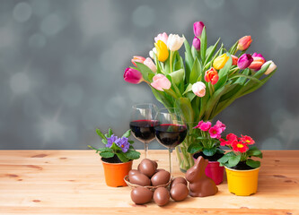 Easter concept. Colorful tulips in vase, chocolate bunny, glasses of red wine, primula flowers and chocolate Easter eggs in wicker basket on wooden table.