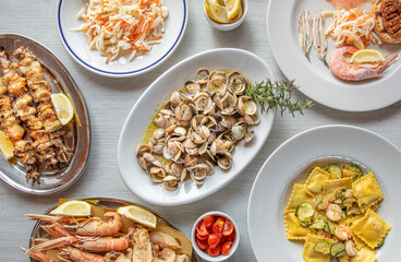 composition of cooked Italian seafood dishes, with fried fish, skewers, clams, anchovies, shrimps and stuffed ravioli , top view with white wooden background